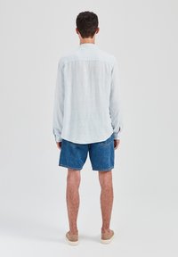 Jasnoniebieska lniana koszula z długimi rękawami, o luźnym kroju i bocznych rozcięciach, zestawiona z dżinsowymi szortami i casualowymi beżowymi butami, widziana od tyłu.