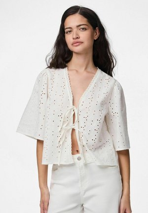 Blouse blanche à manches courtes avec un design à perforations, fermeture à nouer à l'avant et bordure en forme de vagues au niveau du cou. Tissu léger et texturé.