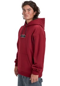 Red fleece hoodie with a front pocket, drawstring hood, and embroidered detailing in green and white. Relaxed fit and soft texture.