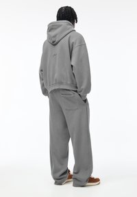 Personne debout, dos à la caméra, portant un sweat à capuche gris ample et un pantalon de jogging assorti, les mains dans les poches, chaussée de baskets marron.