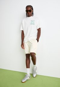 T-shirt blanc à col rond avec logo vert, shorts crème neutres et baskets de sport argentées avec semelle en gomme. Lunettes de soleil noires portées.