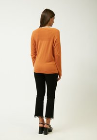 Orange gestrickter Pullover mit strukturierten Details auf dem Rücken, kombiniert mit schwarzen Schlaghosen mit Spitzenkanten am Saum und klobigen Sandalen mit Absatz.