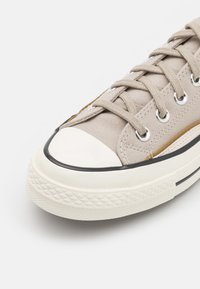 Beige kanvas sneakers med rund vit gummikåpa, svart accentrand, platta snören och metallögon; strukturerad gummisula.