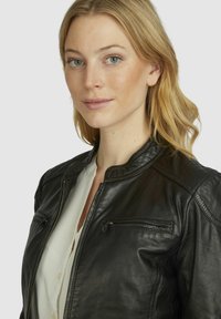 Black leather jacket with a stand collar, quilted shoulder accents, front zip pockets, and smooth texture. Worn over a white blouse.