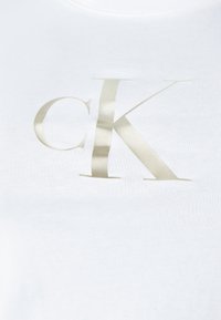 Biała bawełniana koszulka z dużym, metalicznym złotym logo "CK" w nowoczesnej czcionce. Prosty design, gładka tekstura i okrągły dekolt.