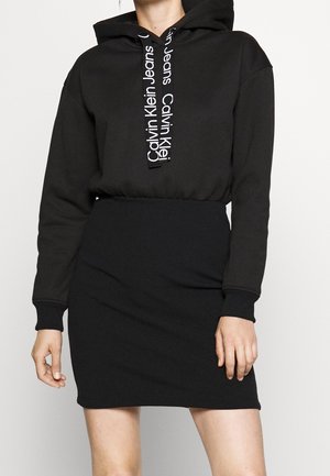 Femme portant une robe à capuche noire avec le logo Calvin Klein Jeans sur les cordons, manches longues et bas de jupe ajusté.