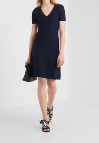 Abito a coste blu navy con maniche corte e scollatura a V, abbinato a una borsa nera e sandali con motivo bianco e nero decorati con fiocchi.