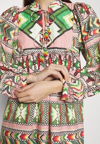 Multikleurige blouse met geometrische patronen in rood, groen, roze en zwart, met poffende mouwen en een keyhole-halslijn met strikjes.