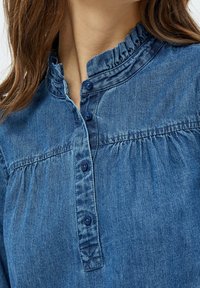 Blue denim shirt featuring a ruffled collar, button-down closure, and subtle gathers at the front. Soft texture with a casual design.