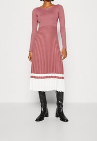 Polven pituinen mauve väriin kuuluva mekko, jossa on ribbikuviointi, pitkät hihat ja valkoinen laskostettu helma, jossa on kaksi mauve-raitaa. Yhdistetty mustiin korkokenkiin.