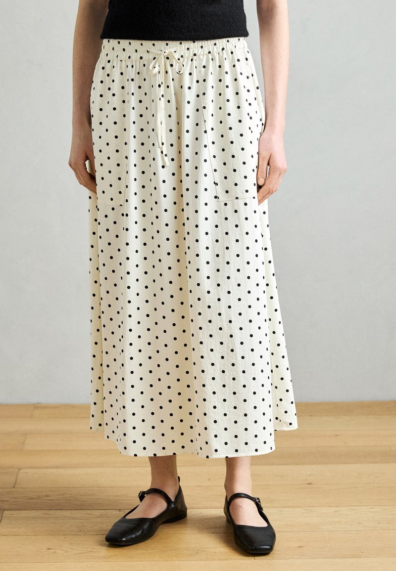 Λευκή φούστα μέχρι το γόνατο με μαύρα πουά, ελαστική μέση με κορδόνι και δύο πλαϊνές τσέπες. Φορεμένη με απλά μαύρα φλατ παπούτσια.