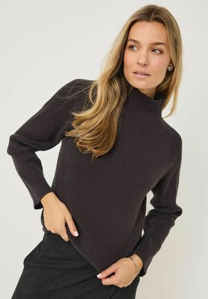 Pull en maille côtelée sombre avec un col montant, des poignets ajustés et une coupe décontractée, porté sur une jupe noire, mettant en valeur des lignes épurées et une texture minimale.