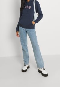 Sweat à capuche bleu marine avec le texte "Ravy", jeans bleu clair à jambes droites, et chaussures à plateforme blanches. Un sac à motif damier complète la tenue.