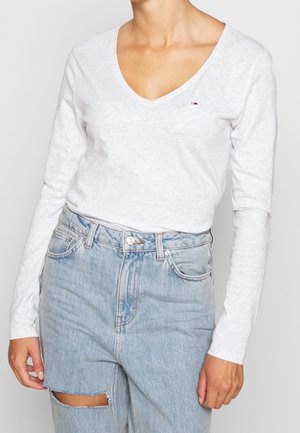 Personne portant un t-shirt blanc à manches longues avec col en V et un petit logo, ainsi qu'un jean déchiré bleu clair taille haute.