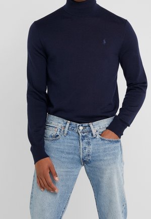 Person trägt einen enganliegenden dunkelblauen Rollkragenpullover mit kleinem Logo auf der Brust und eine hellblaue Jeans, eine Hand in der Tasche.