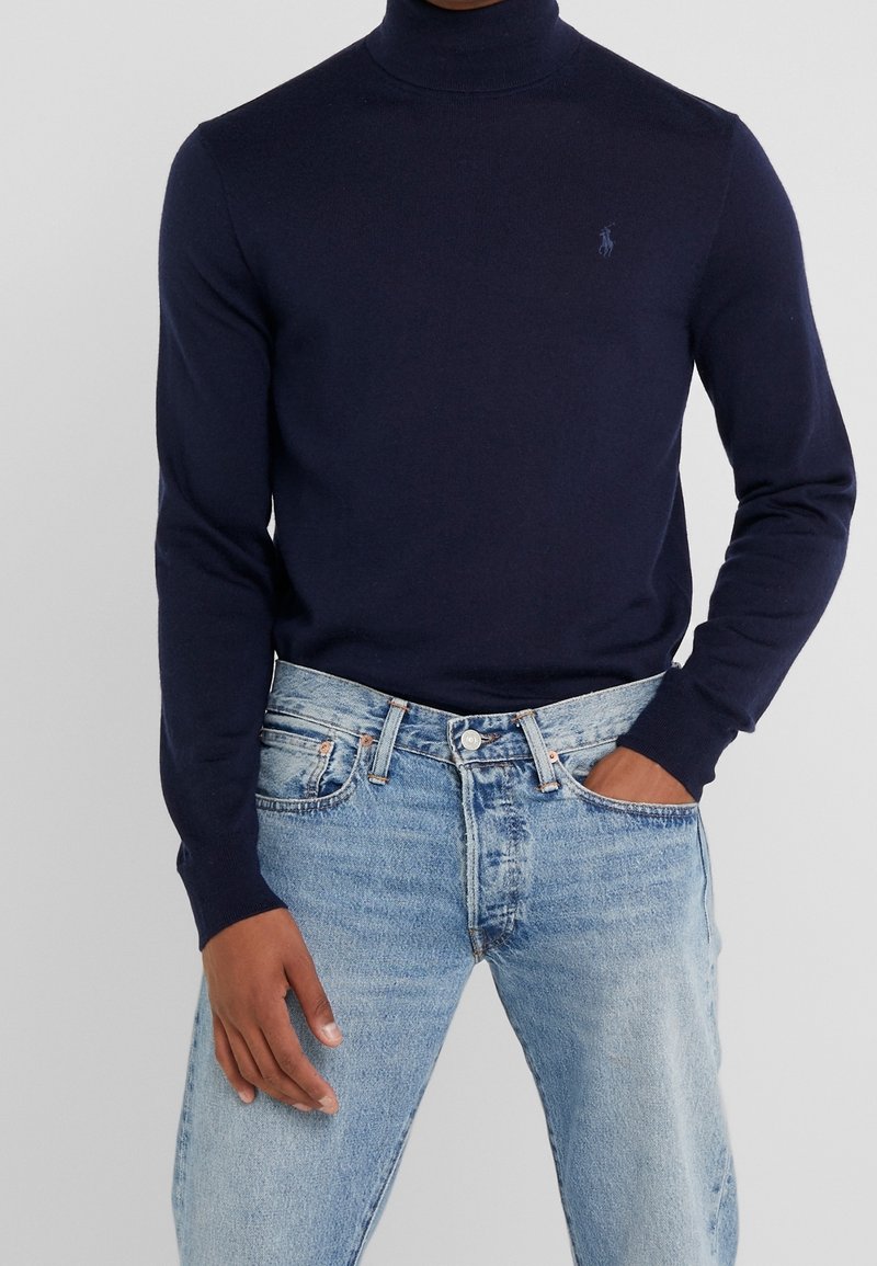 Personne portant un pull à col roulé bleu marine ajusté avec un petit logo sur la poitrine et un jean bleu clair, une main dans la poche.