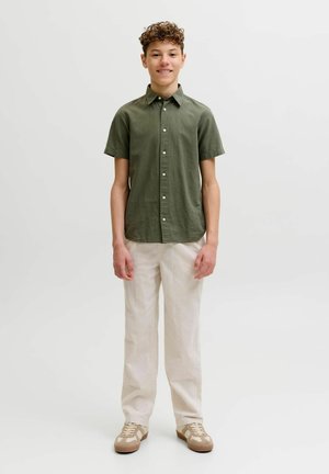 Teenager s kučeravými vlasmi stojí usmievajúc sa, má na sebe olivovo zelenú krátkym rukávom košeľu na gombíky, biele nohavice a béžové tenisky.