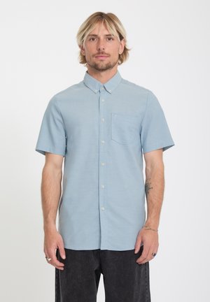 Mann mit blondem Haar, der ein hellblaues kurzärmeliges Hemd mit Knöpfen und dunkle Hose trägt, vor einem schlichten weißen Hintergrund steht.