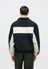 Μπουφάν με μπλοκ χρωμάτων σε μαύρο και ανοιχτό γκρι, με γιακά, ελαστικά μανίκια και σφιχτή μέση. Το ύφασμα φαίνεται λείο και ελαφρύ.