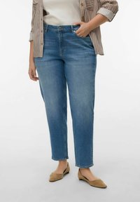 Vrouw die een blauwe spijkerbroek draagt, beige puntige platte schoenen, een wit topje en een beige gestreept overhemd met opgerolde mouwen.