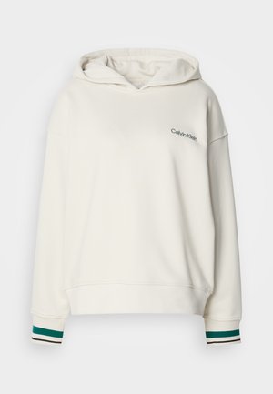 Balta jaku ar kapuci, kurai ir zaļa "Calvin Klein" logo. Piedāvā tumši zaļas ribu piedurknes ar baltām un melnām svītrām. Mīksts, teksturēts audums.