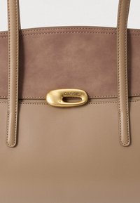 Taupe bőr tote táska, illeszkedő pántokkal és arany ovális Calvin Klein logóval a tetején.