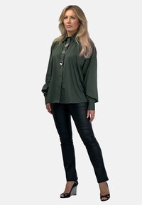 Zielona koszula zapinana na guziki z okrągłym kołnierzykiem i trzema dużymi guzikami, zestawiona z czarnymi dopasowanymi spodniami oraz butami na obcasie z teksturowymi akcentami.