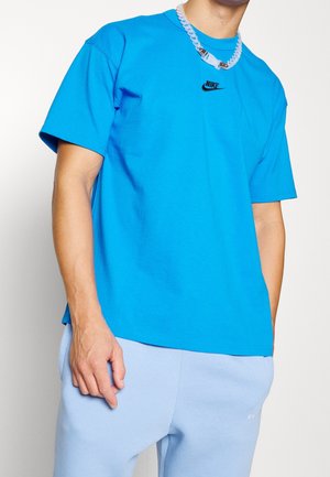 T-shirt in cotone blu con maniche corte, caratterizzato da un logo Nike nero. Abbinato a pantaloni della tuta blu chiaro e a una collana trasparente e voluminoso.