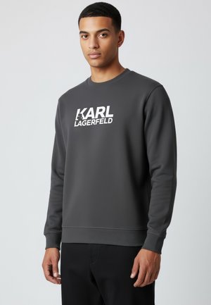 Junger Mann trägt ein dunkelgraues Sweatshirt mit dem weißen Schriftzug "KARL LAGERFELD" auf der Vorderseite, steht vor einem schlichten hellen Hintergrund.