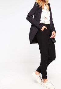Černý dlouhý blazer s kapsami, bílý květinový top pod ním, černé zúžené kalhoty a bílé tenisky, vytvářející vrstvený, pohodlný vzhled.