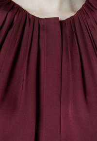 Gros plan d'un tissu bordeaux profond avec des plis verticaux et une couture centrale, drapé autour d'un décolleté nu.