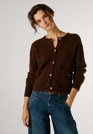 Cardigan marrón de punto con cuello redondo, bolsillos delanteros y botones de madera, combinado con pantalones de mezclilla azul. Textura suave, ajuste relajado.