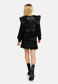 Črna obleka z bleščečim bodijem iz bleščic, nabrano na ramenih in večplastno krilo. Kombinirana s črnimi škornji do kolen. Gladek videz, prilegan kroj.