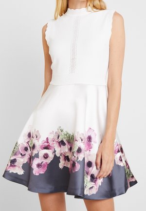 Kobieta w białej sukience na ramiączkach z kwiatowym nadrukiem na dolnej krawędzi spódnicy w odcieniach różu i zieleni, stojąca na jednolitym tle.