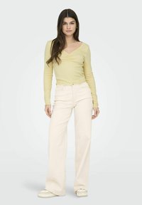 Langslavige, lichtgele top met een V-hals en geplooide details, gecombineerd met hooggeritste, crèmekleurige wijde jeans en witte sneakers.