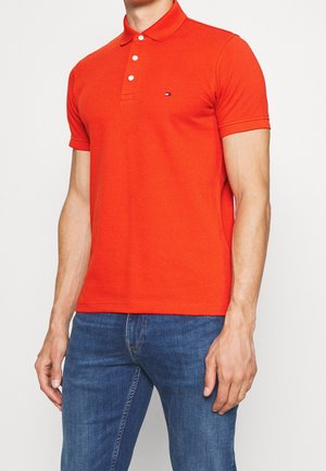 Homme portant un polo à manches courtes orange vif avec trois boutons blancs et un jean bleu, debout devant un fond blanc uni.