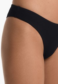 Černé bikini kalhotky z texturované látky. Mají nízký pas a minimální zakrytí, které zvýrazňuje hladký tón pleti.