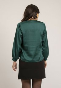 Blouse en satin vert foncé avec des manches bouffantes, un col rond et un ourlet incurvé, portée sur une jupe noire, mettant en valeur la texture lisse.