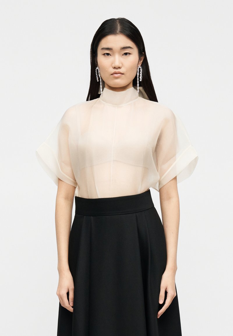 Femme aux longs cheveux foncés portant un chemisier beige transparent, une jupe taille haute noire, et de longues boucles d'oreilles pendantes en cristal, sur un fond uni.