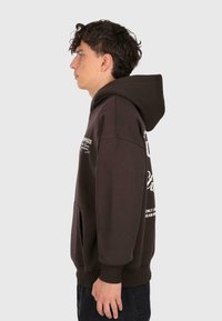 Jeune homme de profil portant un sweat à capuche marron foncé avec du texte blanc, se tenant contre un fond gris clair uni.