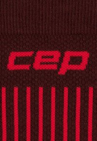Kompressionssocken in Dunkelbraun mit roten Akzenten. Sie verfügen über eine strukturierte Strickoberfläche und das "cep"-Logo, das auffällig in leuchtendem Rot präsentiert wird.