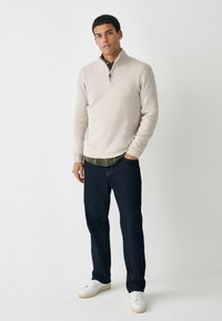 Pull à col zippé beige sur une chemise vert foncé, assorti à un jean en denim noir et des baskets blanches. Texture lisse, coupe décontractée.