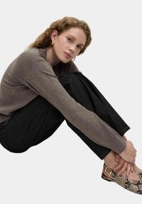 Maglione marrone a maglia, pantaloni neri a gamba larga e ballerine con motivo serpente. Modella seduta, mani incrociate, contro uno sfondo neutro.