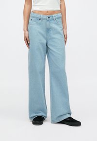 Άτομο που φοράει ανοιχτό μπλε τζιν παντελόνια φαρδιάς γραμμής, μαύρα αθλητικά παπούτσια και λευκό crop top αμάνικο, στέκεται μπροστά σε απλό φόντο.