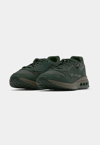 Sapatilhas Reebok verde-escuro com painéis de camurça texturizada e malha, atacadores pretos e uma sola espessa com recortes circulares sobre um fundo branco.