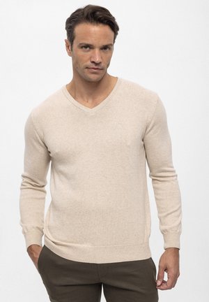 Jersey beige de cuello en V con mangas largas, confeccionado en material suave, con puños y dobladillo acanalados, usado sobre pantalones oscuros.