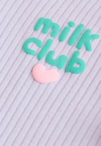 Mehke zelene črke, ki sestavljajo "milk club" nad rožnim srcem na svetlo vijolični rebrasti tkanini.