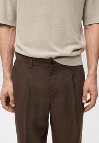 Pull en maille beige associé à un pantalon marron. Le pantalon présente une fermeture à boutons et des plis à l'avant, mettant en valeur une texture de tissu lisse.