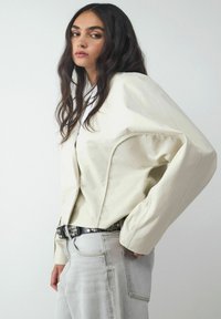 Kremowa kurtka z sztucznej skóry o oversize'owym kroju, kontrastowym przeszyciu i zakrzywionym końcu, w połączeniu z jasnoszarymi dżinsami i dekoracyjnym czarnym paskiem.