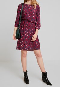 Krátký rukáv šaty z tmavě burgundské látky s hravým puntíkovým vzorem v červené, růžové a modré; úzký pas a volný střih; kombinováno s černými kotníkovými botami.